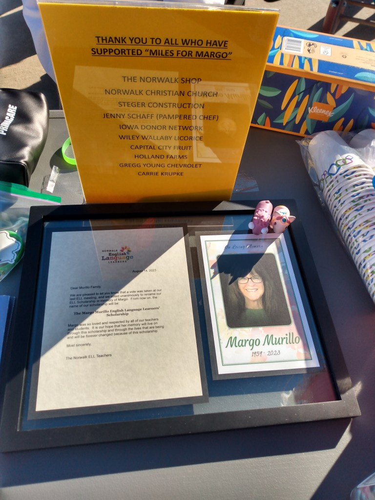 Two toy unicorns sitting on a framed memorial that reads: 
August 14, 2023
Dear Murillo Family
We are pleased to let you know that a vote was taken at our last ELL meeting, and we voted unanimously to rename our ELL Scholarship in memory of Margo. From now on, the name of our scholarship will be:
The Margo Murillo English Language Learners' Scholarship
Margo was so loved and respected by all of our teachers and students. It is our hope that her memory will live on through this scholarship and through the lives that are being and will be forever changed because of this scholarship
Most sincerely,
The Norwalk ELL Teachers
In loving memory of Margo Murillo, 1959-2023

There is a donor list behind them that reads:
THANK YOU TO ALL WHO HAVE SUPPORTED "MILES FOR MARGO"
THE NORWALK SHOP
NORWALK CHRISTIAN CHURCH 
STEGER CONSTRUCTION 
JENNY SCHAFF (PAMPERED CHEF) 
CARRIE KRUPKE
IOWA DONOR NETWORK
WILEY WALLABY LICORICE
CAPITAL CITY FRUIT 
HOLLAND FARMS
GREGG YOUNG CHEVROLET